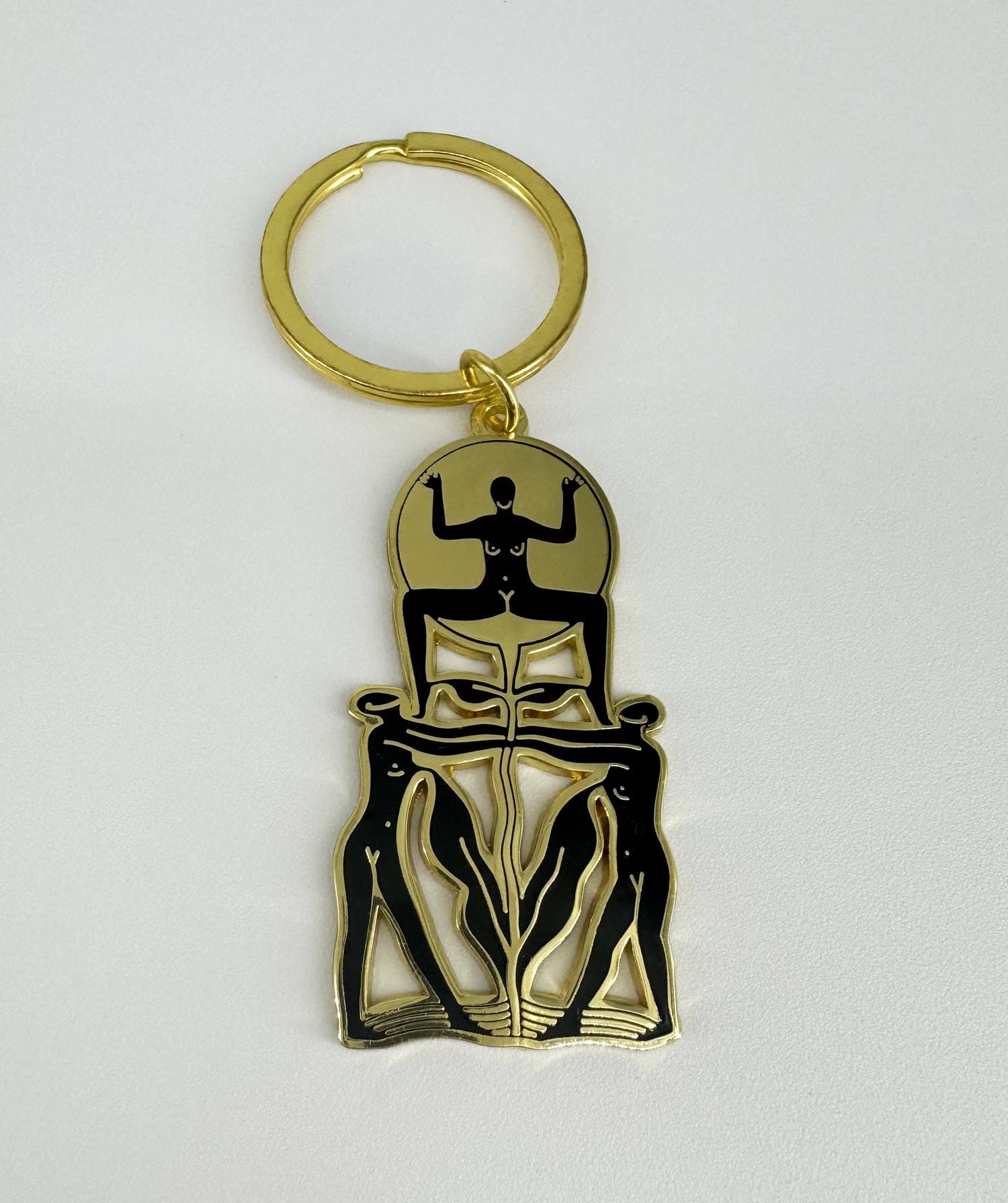 A golden keychain with a hard enamel design featuring a stylized, black-outlined figure. The keychain has a four-link chain and a keyring.