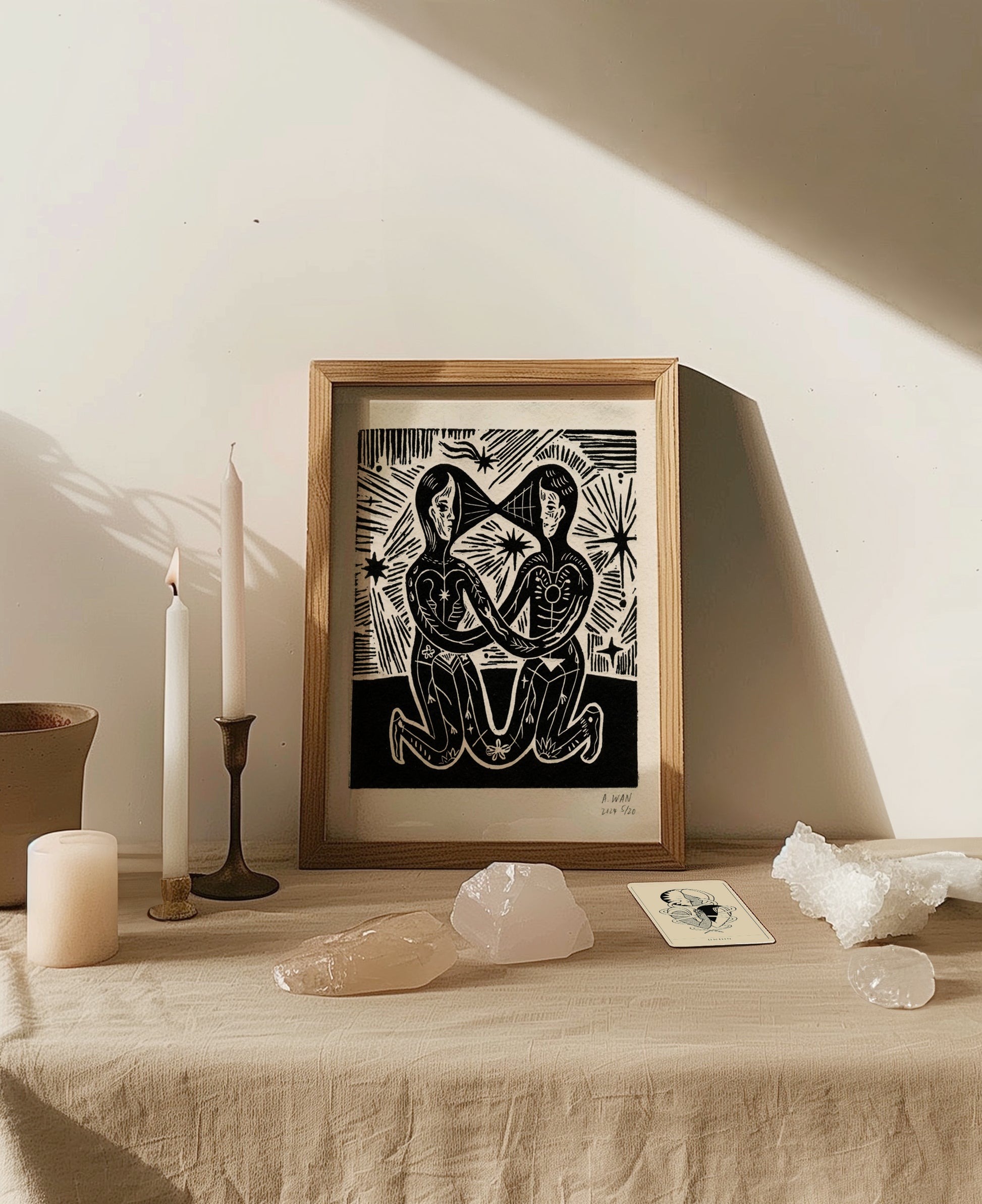 Linocut print titled Union by Andrea Wan, depicting two mirrored human figures with botanical and cosmic markings, kneeling face to face, hands intertwined, against a backdrop of stars and energy lines. Printed in black ink on textured paper.

